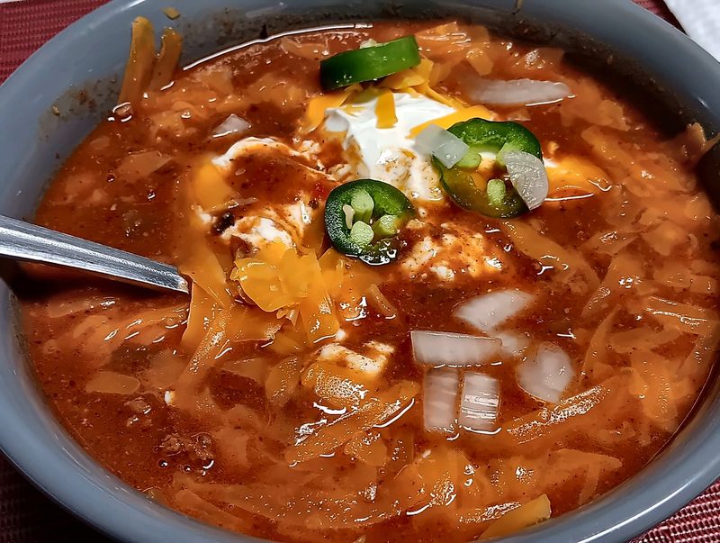 A bowl of chili topped with shredded cheddar, sour cream, jalapeño slices and onion